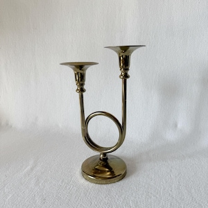 Puede incluir: Un candelabro de latón con dos copas para velas. El diseño presenta una forma curva, similar a un cuerno, que conecta las copas a una base circular. El metal tiene un acabado pulido, que refleja la luz. Es un artículo decorativo.