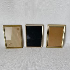 Può includere: Tre cornici rettangolari con bordi in metallo color oro. Le cornici hanno sfondi di diversi colori: uno con sfondo beige chiaro, uno con sfondo nero e uno con sfondo beige. Le cornici sono disposte orizzontalmente.
