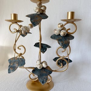 Puede incluir: Un candelabro de metal dorado con tres candeleros. El candelabro presenta hojas decorativas con detalles en azul y dorado y racimos de bayas blancas y doradas. La base es redonda y dorada.
