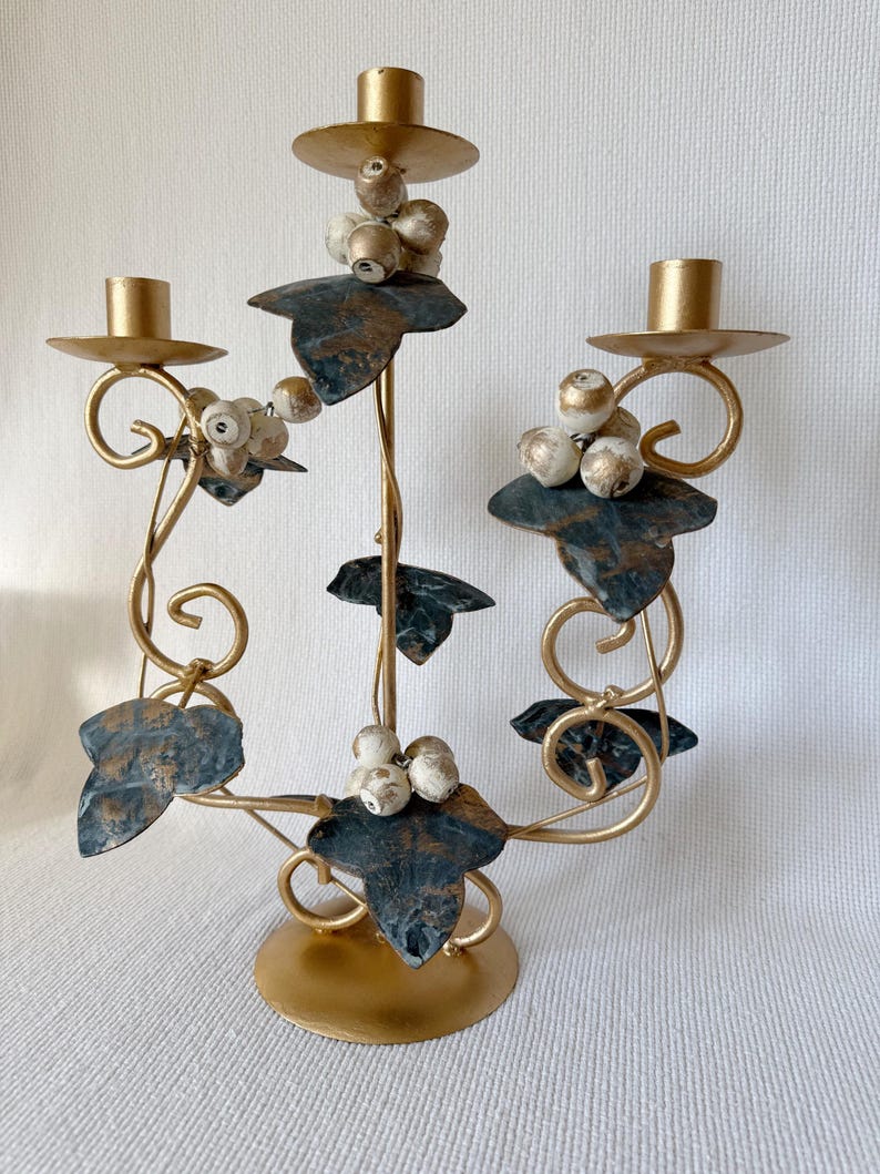 Puede incluir: Un candelabro de metal dorado con tres candeleros. El candelabro presenta hojas decorativas con detalles en azul y dorado y racimos de bayas blancas y doradas. La base es redonda y dorada.