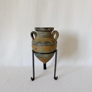 Op de afbeelding: Een decoratieve keramische vaas in antieke stijl, met twee handvatten, in bruin, groen en beige tinten. De vaas is versierd met geometrische patronen en staat op een zwarte metalen standaard met drie poten.