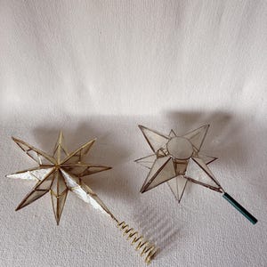 May include: Two star-shaped ornaments constructed from translucent white panels framed in gold-toned metal. One star features a gold-toned metal coil base for mounting, while the other star includes a straight dark green stem attachment.