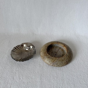 May include: Two decorative items: a silver-colored, nautilus-shaped dish and a round, beige stone bowl. The nautilus dish has a detailed, ridged design. The stone bowl has a smooth, polished surface and a circular opening.