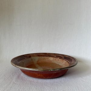 Puede incluir: Un cuenco de cerámica hecho a mano con un diseño ancho y poco profundo. El cuenco presenta un exterior marrón rojizo y un interior más claro con una superficie texturizada. El borde tiene un patrón decorativo de líneas marrones.