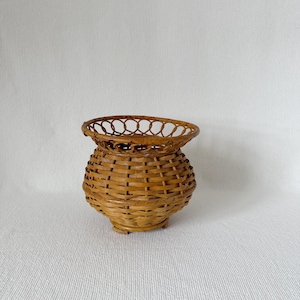May include: A small, round, woven basket with a flared top and a decorative openwork rim. The basket is made of light brown wicker and has a bulbous body. It sits on three small feet.