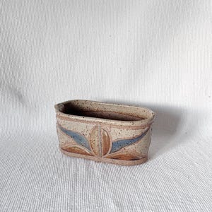 May include: A small, rectangular ceramic planter with a speckled beige glaze. The planter has a decorative design of stylized leaves in shades of blue, brown, and beige.