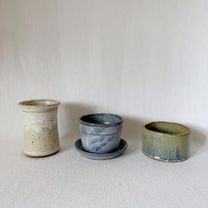 May include: Three handmade ceramic planters in neutral tones. The tallest is a beige cylinder with a textured pattern. The middle planter is blue-grey with a matching saucer. The third is a low, wide planter with green and blue glaze.