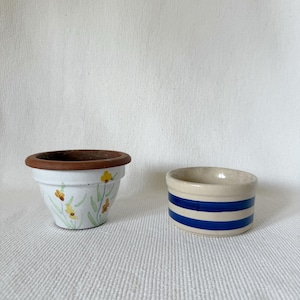 Op de afbeelding: Twee kleine keramische potten. Eén is wit met een bruine rand en een geel bloemmotief. De andere is een kleine, beige container met twee blauwe horizontale strepen.