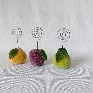 May include: Three fruit-shaped place card holders featuring a yellow citrus, a purple plum, and a green pear. Each piece has a textured, sugar-like finish, a single green leaf, and a silver-colored metal wire spiral extending from the top.