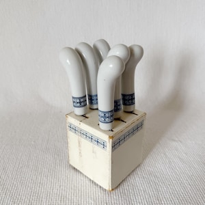 Puede incluir: Un juego de seis mangos de cuchillo de cerámica blanca con diseños florales azules, dispuestos en una caja de madera color crema. Los mangos tienen forma curva y se exhiben sobre un fondo blanco.