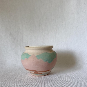 Könnte beinhalten: Eine kleine, runde Keramikvase mit cremefarbenem Oberteil und rosa Körper. Die Vase ist mit einem bergähnlichen Design in Grün- und Brauntönen verziert. Die Vase hat eine kleine Öffnung oben.