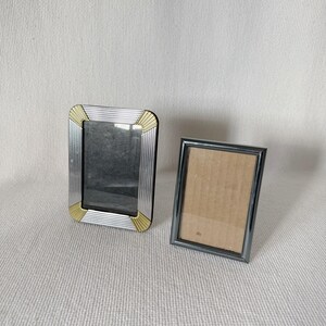 May include: Two silver picture frames, one with a gold trim, on a white textured surface.