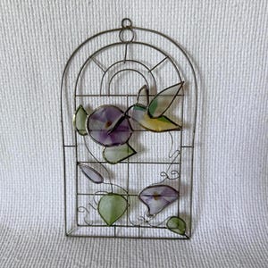 Puede incluir: Un panel de ventana de vidrieras con un colibrí y glorias de la mañana. El marco de metal tiene forma de arco con un diseño de rejilla. El colibrí tiene detalles amarillos, verdes y blancos. Las flores son moradas y las hojas son verdes.