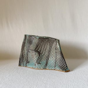 Puede incluir: Un jarrón de cerámica hecho a mano con una superficie texturizada única. El jarrón tiene una forma triangular con un patrón moteado en tonos grises y verde azulado. La base tiene un acabado natural sin esmaltar.