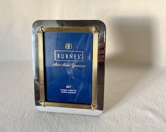 May include: A rectangular silver-toned picture frame with gold-colored accents. The frame has a blue interior and features the brand name "BURNES" and the size "5x7". The frame is lacquer coated for tarnish resistance.