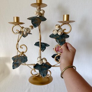Puede incluir: Un candelabro de metal dorado con tres candeleros. El candelabro presenta hojas decorativas y racimos de cuentas blancas y doradas. Las hojas son de color verde oscuro con un acabado envejecido.