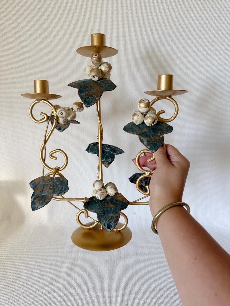 Puede incluir: Un candelabro de metal dorado con tres candeleros. El candelabro presenta hojas decorativas y racimos de cuentas blancas y doradas. Las hojas son de color verde oscuro con un acabado envejecido.