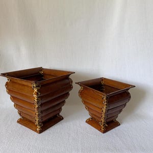 Peut inclure: Deux jardinières carrées en bois. Les jardinières sont en bois brun foncé et sont cousues ensemble avec un matériau de couleur plus claire. La plus grande jardinière mesure environ 25 cm de haut, et la plus petite environ 18 cm.