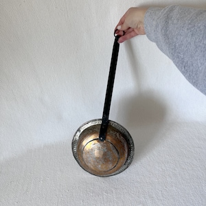 May include: A copper ladle with a black metal handle. The ladle has a rounded bowl and a decorative rim.