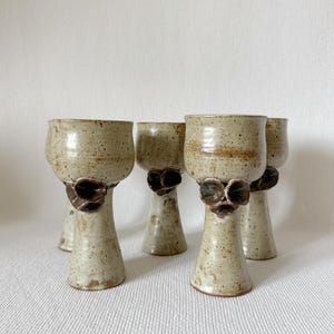 Puede incluir: Conjunto de cinco copas de cerámica hechas a mano. Acabado beige moteado con un adorno floral marrón oscuro en la base del cuenco. Los tallos son acampanados y los cuencos anchos. Miden unos 15 cm de alto.
