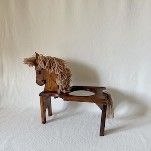 Könnte beinhalten: Ein hölzerner, pferdeförmiger Hocker mit dunkelbraunem Finish. Das Pferd hat eine Mähne und einen Schweif aus beigefarbenem Garn. Der Hocker hat eine kreisförmige Öffnung im Sitz und vier Beine. Der Pferdekopf ist mit einem Zaumzeug versehen.