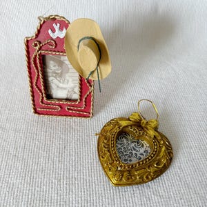 May include: A red and gold picture frame with a small cowboy hat and a gold heart-shaped frame. The red frame has a picture of a child in a cowboy hat. The gold heart frame has a decorative design.