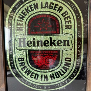 Puede incluir: Un letrero enmarcado de Cerveza Lager Heineken. La etiqueta verde y dorada presenta el logotipo de Heineken, texto y una estrella roja. El letrero dice "Brewed in Holland" y "12 fl oz". El marco es de madera.