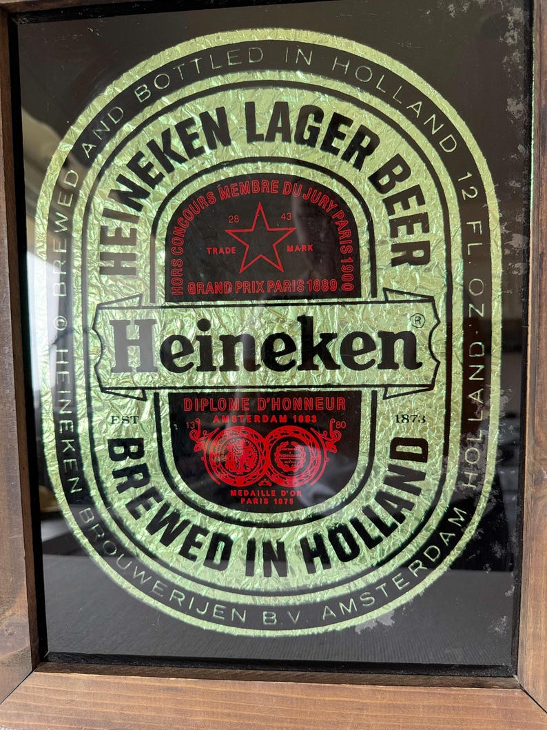 Puede incluir: Un letrero enmarcado de Cerveza Lager Heineken. La etiqueta verde y dorada presenta el logotipo de Heineken, texto y una estrella roja. El letrero dice "Brewed in Holland" y "12 fl oz". El marco es de madera.