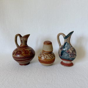 May include: Three small, hand-painted ceramic pitchers. The pitchers are in shades of brown, beige, and red, with floral and geometric designs. Each pitcher has a handle and a spout. The background is a white, textured surface.