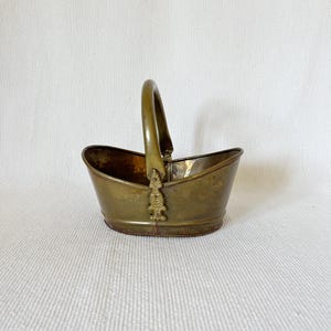 May include: An antique brass basket with a curved handle. The basket has an oval base and a decorative floral design on the handle attachment. The brass has a warm, aged patina, suggesting its vintage nature. The basket is empty.