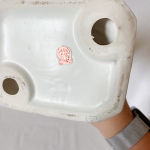 May include: The bottom of a white ceramic figurine with a red and white sticker that reads "Ryuko Made in Japan".