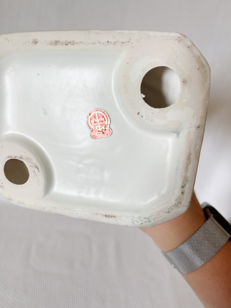 May include: The bottom of a white ceramic figurine with a red and white sticker that reads "Ryuko Made in Japan".