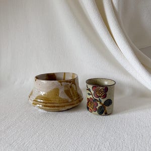 May include: Two ceramic items are displayed: a wide, shallow bowl with a tan and brown glaze and a small, cylindrical cup adorned with red and yellow floral patterns on a beige base. They are set against a white background.