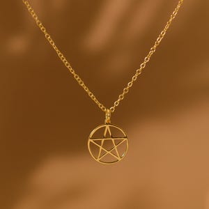 May include: A gold-coloured necklace with a pendant featuring a pentagram design inside a circular frame. The chain and pendant have a warm gold tone, set against a blurred brown background. The necklace is a simple, elegant piece of jewellery.