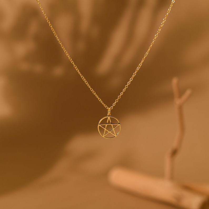 May include: A gold-colored necklace featuring a pendant with a pentagram design within a circular frame. The chain and pendant are a warm gold tone, set against a blurred brown background. The necklace is a simple, elegant piece of jewelry.
