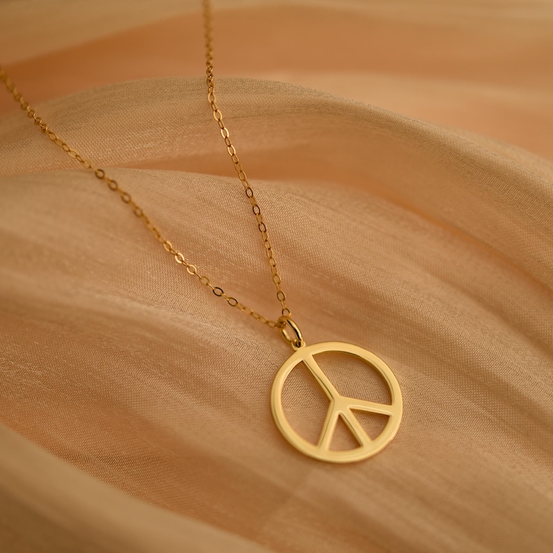 May include: A gold-colored necklace featuring a peace symbol pendant. The pendant is a circle with a three-pronged design inside. The necklace chain is delicate and hangs on a peach-colored fabric.
