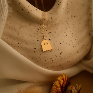 Może przedstawiać: Złoty naszyjnik z wisiorkiem w kształcie ducha. Wisiorek ma prosty, stylizowany kształt ducha z dwoma małymi otworami na oczy. Łańcuszek naszyjnika jest delikatny i złoty. Tło jest w neutralnym kolorze z teksturowaną powierzchnią.