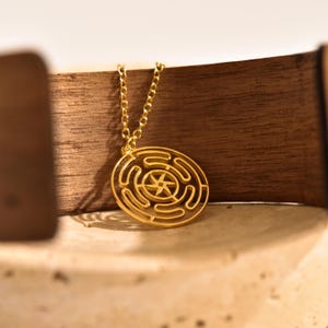 May include: A gold-colored pendant necklace featuring a circular labyrinth design with a star in the center. The pendant hangs from a delicate chain. The necklace is displayed against a wooden background.