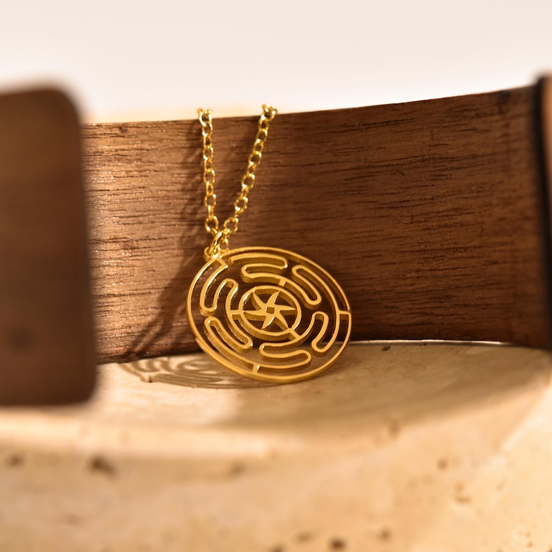 May include: A gold-colored pendant necklace featuring a circular labyrinth design with a star in the center. The pendant hangs from a delicate chain. The necklace is displayed against a wooden background.