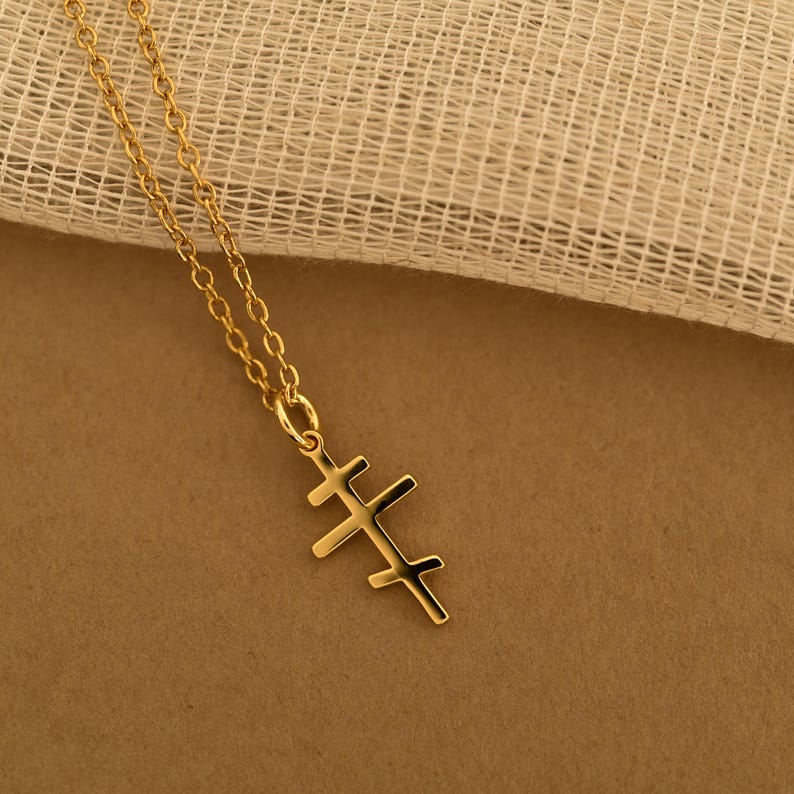 May include: A gold-colored necklace with a pendant featuring a triple cross design. The chain and pendant are made of a shiny, reflective metal. The necklace is displayed against a neutral-toned background.