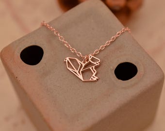 Peut inclure: Un collier en or rose avec un pendentif délicat en forme d'écureuil origami. Le collier est posé sur une surface texturée brune.