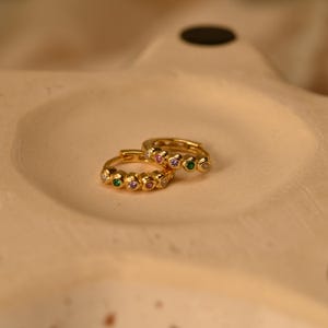 May include: Two gold-colored hoop earrings, each adorned with a series of small, colorful gemstones. The gemstones include green, purple, pink, and clear stones. The earrings are displayed on a light beige surface.