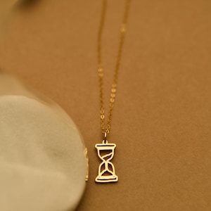 May include: A gold-colored necklace featuring an hourglass pendant. The pendant is a detailed cutout of an hourglass shape. The chain is delicate and hangs from the pendant. The background is a warm, neutral tone.