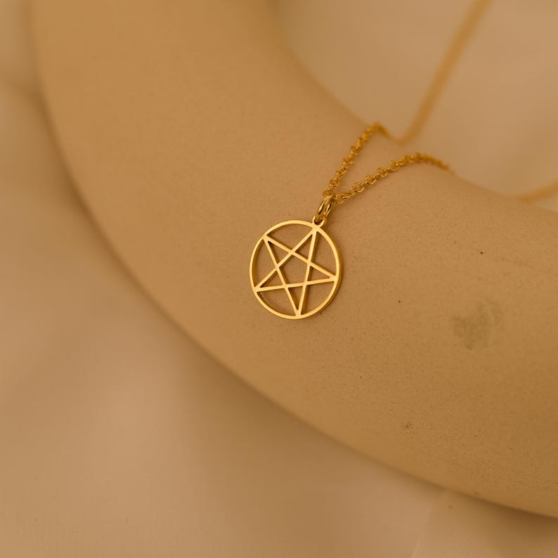 May include: A gold-colored pendant necklace featuring a pentagram design within a circular frame. The necklace chain and pendant are a matching gold tone. The pendant is centered in the image, with a soft, neutral background.