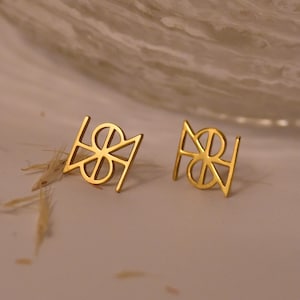 May include: A pair of gold-colored stud earrings. Each earring features a geometric design with a central circular element and angular lines. The earrings are displayed on a light-colored surface, with a blurred background.