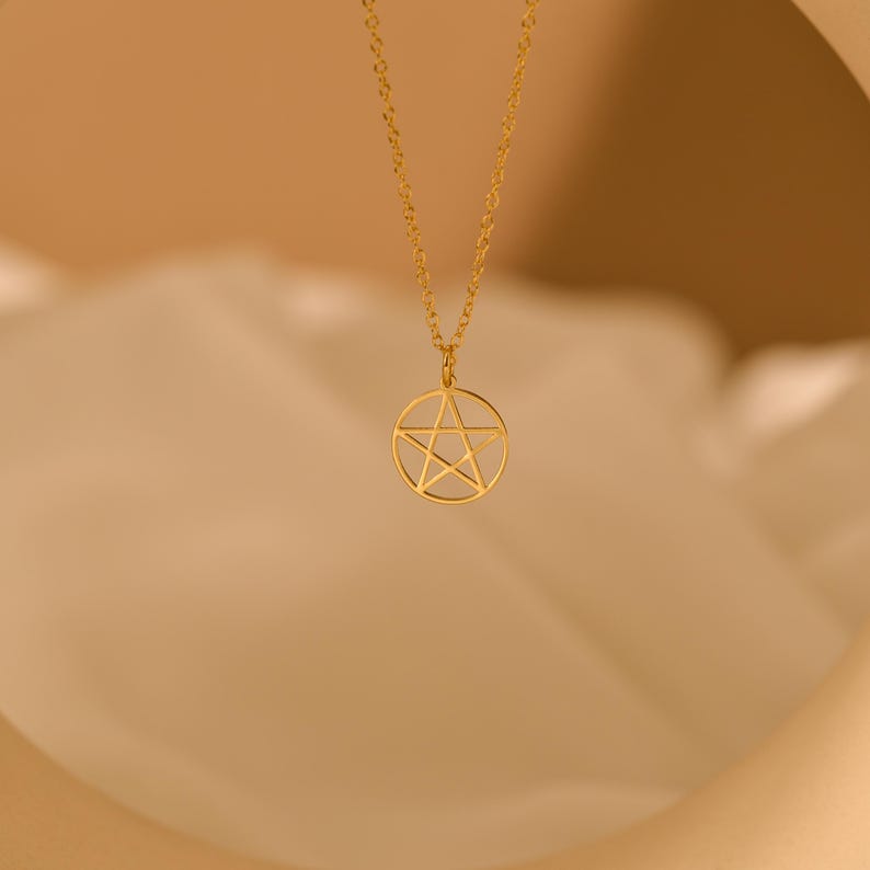 May include: A gold-colored pendant necklace featuring a pentagram design within a circle. The necklace has a delicate chain and a simple, elegant aesthetic. The pendant is suspended from the chain, creating a focal point. The background is a soft, neutral color.