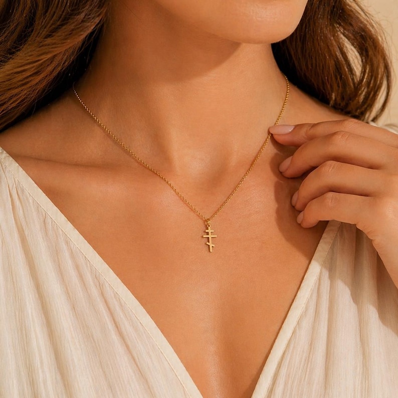 May include: A gold-colored necklace with a pendant in the shape of a cross. The necklace is worn around the neck. The pendant has a unique design with multiple horizontal bars. The background is a soft, neutral color.