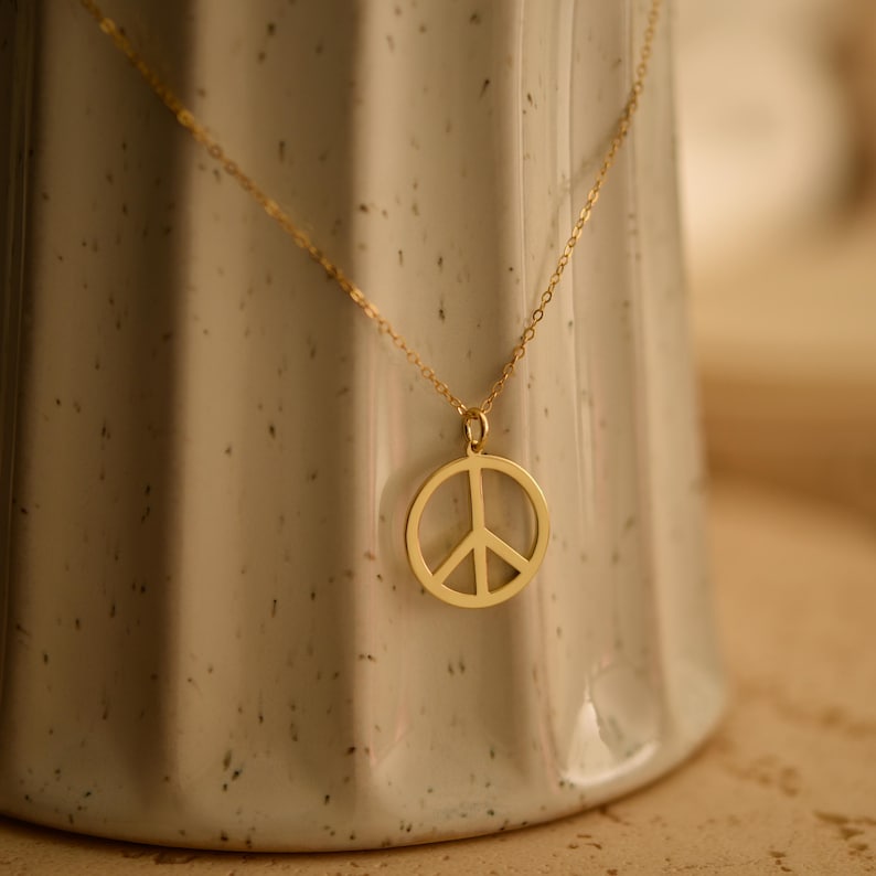 May include: A gold-colored necklace featuring a peace symbol pendant. The pendant is a circular design with a vertical line bisected by two diagonal lines. The necklace is displayed against a neutral background.