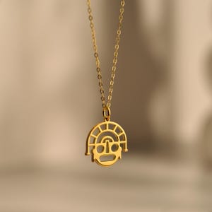 Op de afbeelding: Een goudkleurige hangerketting met een gestileerd gezichtsontwerp. De hanger is rond met een gezicht-achtige vorm en een zon-achtig ontwerp erboven. De ketting is delicaat en goudkleurig, wat een eenvoudig maar elegant accessoire creëert.