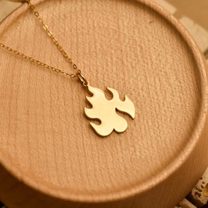 May include: A gold-colored necklace with a flame-shaped pendant. The pendant is smooth and shiny, and the chain is delicate. The necklace is displayed on a wooden surface.